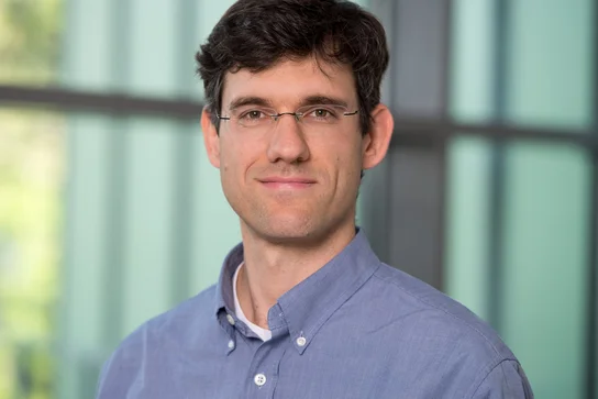 Ein Mann mit braunem Haar und einer Brille trägt ein hellblaues Hemd. Er steht vor einer Fensterfront mit unscharfem Hintergrund und lächelt leicht.