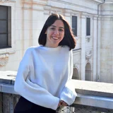 Eine junge Frau mit langen, dunklen Haaren trägt einen weißen Pulli und steht lächelnd an einem Geländer. Im Hintergrund sind historische Gebäude mit architektonischen Details zu sehen. Die Szene vermittelt ein Gefühl von Freude und Anmut.