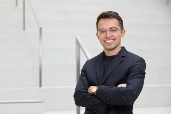 Ein junger Mann in einem schwarzen Blazer steht an einer Treppe, lächelt freundlich und hat die Arme verschränkt. Er trägt eine Brille und wirkt selbstbewusst. Die Umgebung ist modern und hell.