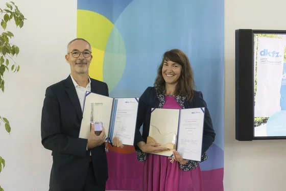 Zwei Personen stehen nebeneinander, beide lächeln. Der Mann trägt einen dunklen Anzug und hält einen Pokal sowie Dokumente in der Hand. Die Frau trägt ein pinkes Kleid und hält ebenfalls Dokumente.