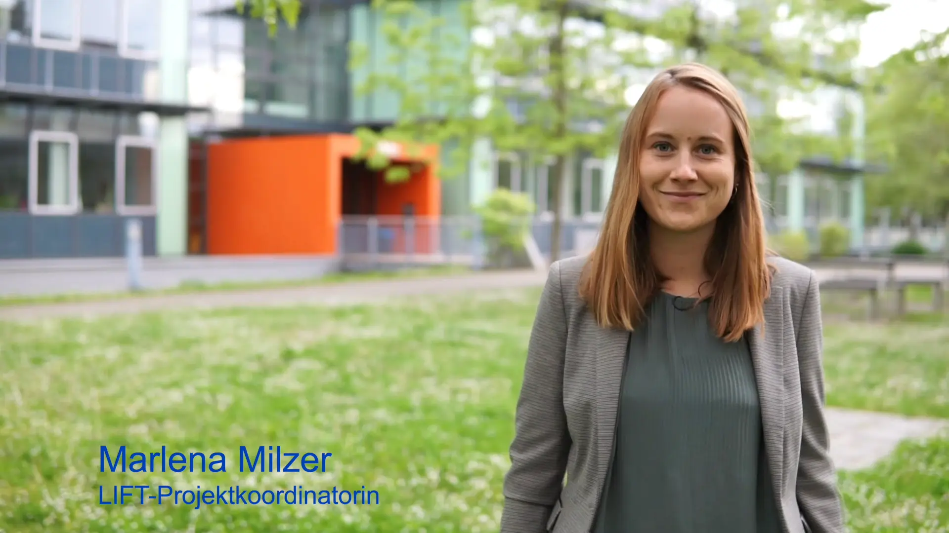 Marlena Milzer steht im Freien vor einem modernen Gebäude mit Glasfassade. Sie trägt ein graues Blazer und lächelt. Neben ihr steht ihr Name und Titel: "LIFT: Projektkoordinatorin". Im Hintergrund sind grüne Wiesen und Bäume sichtbar.