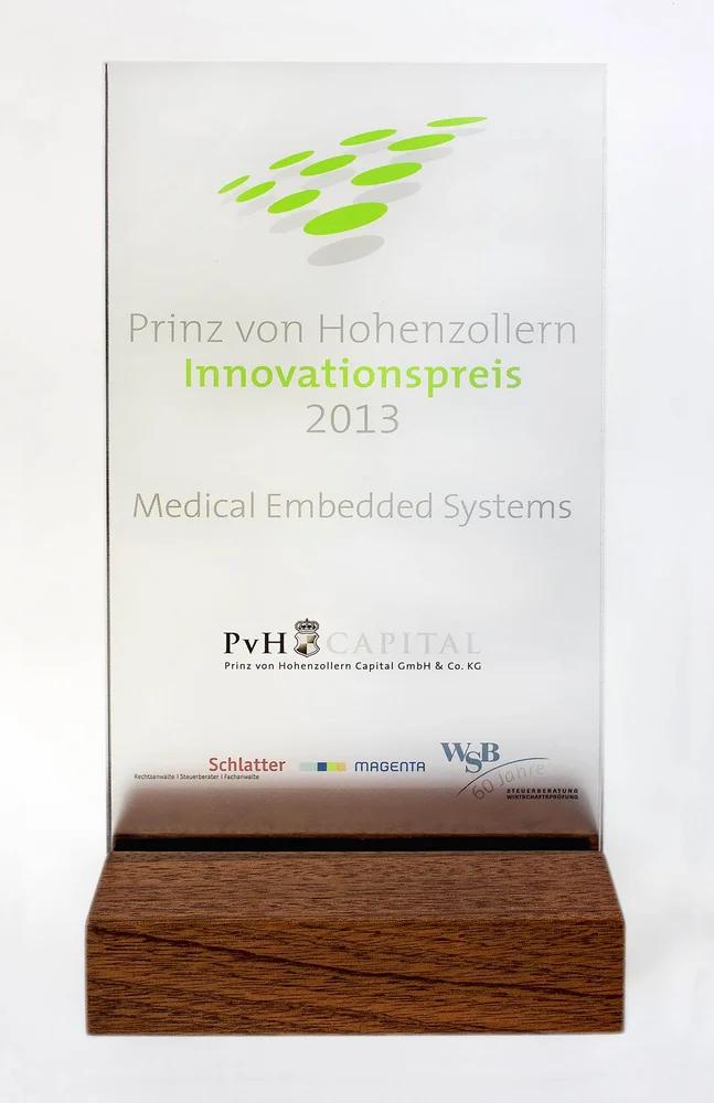 Transparent award trophy featuring the engraved text "Prinz von Hohenzollern Innovationspreis 2013" at the top, and "Medical Embedded Systems" below. The trophy rests on a wooden base and reflects a modern, professional design, symbolizing recognition in the field of innovation.