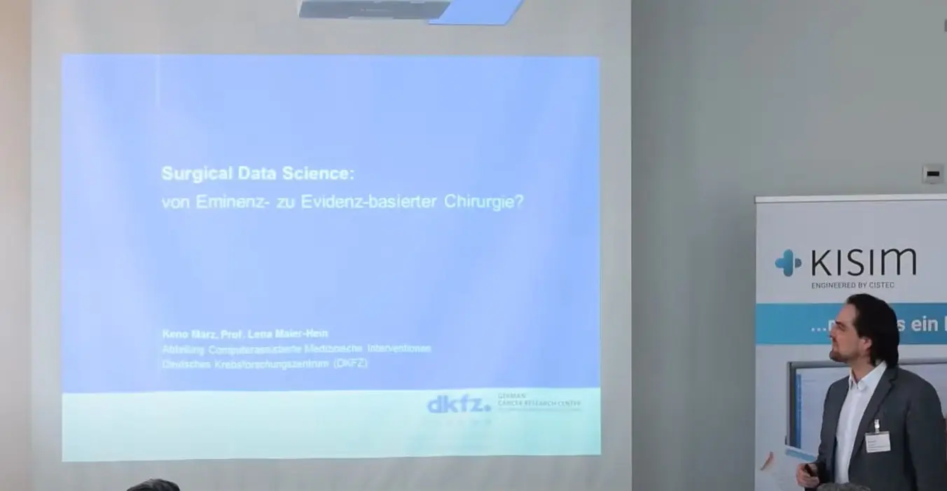 A speaker presents a slide titled "Surgical Data Science: von Eminenz- zu Evidenz-basierter Chirurgie?" in a professional setting. The slide lists the speaker's name and affiliation with dkfz, suggesting a discussion on the intersection of data science and evidence-based surgery.