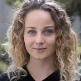 Ein Porträt einer Frau mit langen, lockigen, blonden Haaren und blauen Augen. Sie trägt ein schwarzes Oberteil und hat einen neutralen, freundlichen Gesichtsausdruck. Hintergrund unscharf mit grünen Pflanzen.