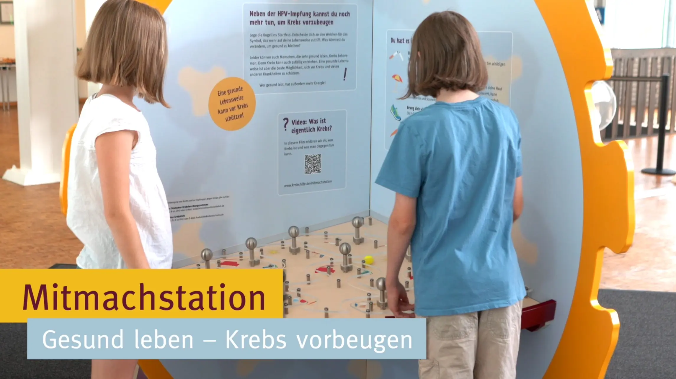 Zwei Kinder stehen an einer interaktiven Station, die sich mit gesundem Leben und Krebsprävention beschäftigt. Die Station gibt Informationen und Anregungen, wie man gesund leben kann, um Krankheiten vorzubeugen.