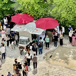Eine große Menschenmenge steht um einen Food Truck mit roten Sonnenschirmen. Die Szene ist von üppigem Grün umgeben, und viele Personen sind in Gespräche vertieft oder warten in der Schlange.