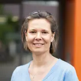 Eine Frau lächelt in die Kamera. Sie trägt ein hellblaues Oberteil und eine dezente Halskette. Ihr Haar ist schulterlang und leicht gewellt. Im Hintergrund sind unscharfe, grüne Elemente und eine orangefarbene Wand sichtbar.