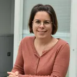 Eine Frau mit braunem Haar, die eine rosafarbene Strickjacke trägt, lächelt und lehnt an einem Geländer. Sie trägt Brille und hat die Hände gefaltet. Der Hintergrund zeigt Fenster und eine helle Innenraum-Umgebung.