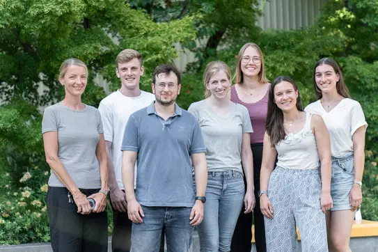 Eine Gruppe von sieben Personen steht lächelnd im Freien, umgeben von Pflanzen. Sie tragen lässige Kleidung und scheinen eine freundliche Atmosphäre zu genießen. Die Gruppe besteht aus drei Männern und vier Frauen.
