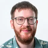 Ein Mann mit Brille und braunem Bart lächelt in die Kamera. Er trägt ein gemustertes Hemd mit floralen Motiven. Der Hintergrund ist einfarbig und hell.