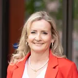 Eine Frau mit long blonden Haaren steht lächelnd an einem Geländer, gekleidet in einem roten Blazer und roten Hosen. Sie trägt ein weißes T-Shirt und sportliche Schuhe. Im Hintergrund sind unscharf Bäume und Gebäude zu sehen.