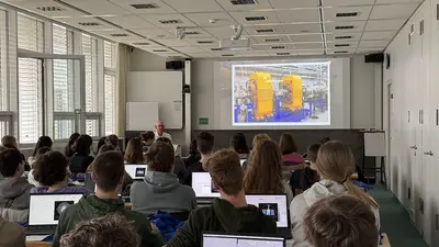 Ein Dozent spricht vor einer Gruppe von Studierenden in einem Klassenraum. Auf einer Präsentationsleinwand ist ein Bild von Maschinen in einer Industrieumgebung zu sehen. Die Studierenden sind mit Laptops beschäftigt und hören dem Vortrag aufmerksam zu.