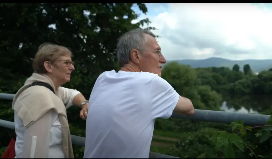 Älteres Paar steht an einem Geländer und blickt nachdenklich in grüne Landschaft.
