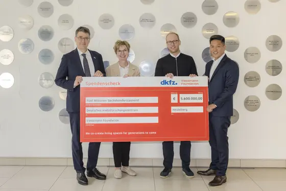 Zwei Männer und zwei Frauen stehen zusammen und halten einen großen Spendencheck über 500.000 Euro für die DKFZ Viessmann Foundation. Im Hintergrund sind Wandplatten mit verschiedenen Namen zu sehen. Der Check ist rot mit der Aufschrift „Spendencheck“ und weiteren Details zur Spende.