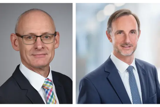 Zwei Porträtfotos von Männern in formeller Kleidung. Der linke Mann trägt eine Brille und ein gestreiftes, mehrfarbiges Krawatte. Der rechte Mann hat einen gepflegten Bart und trägt eine dunkelblaue Jacke mit einer weißen Hemd und einer gepunkteten Krawatte. Beide lächeln leicht.