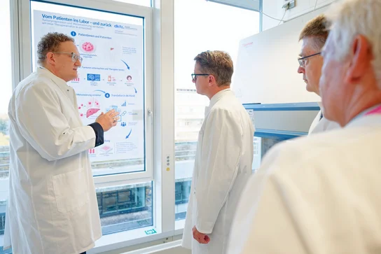 Eine Gruppe von vier Personen in weißen Kitteln steht im Labor. Ein Mann zeigt auf ein Informationsplakat an der Wand. Die Gruppe hört aufmerksam zu und diskutiert.