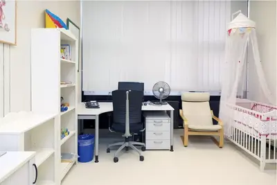 A bright, organized room features a desk with a computer, an office chair, and a fan. To the right, a cozy nursing chair is positioned next to a crib adorned with a canopy. A white bookshelf holds various items, completing the room's functional and inviting atmosphere.