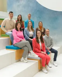 A diverse group of nine trainees sits on colorful steps in an indoor setting, smiling confidently. The trainees are casually dressed and display a camaraderie, reflecting a vibrant and supportive atmosphere related to their administration program.