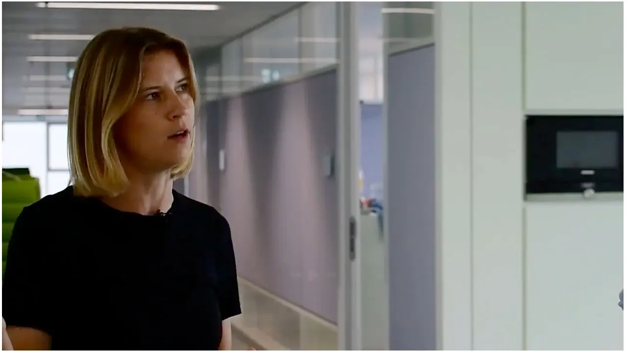 A woman with shoulder-length blonde hair is speaking in an office setting. She is wearing a black shirt and appears engaged in conversation, conveying a sense of professionalism and focus. The background features modern office decor.