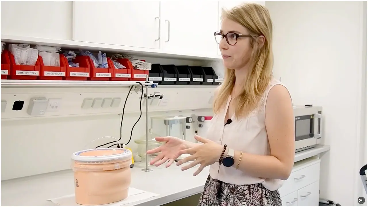 A woman with blonde hair and glasses stands in a laboratory, explaining a process. In front of her is a container with a lid, which appears to hold a substance related to her discussion. The lab features shelves with organized materials in the background.