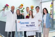 Eine Gruppe von sechs Personen in weißen Laborkitteln steht zusammen und hält Blumen sowie ein großes Schild mit der Aufschrift "96.460€". Darin wird auf eine Spende für eine Kindheit ohne Krebs hingewiesen. Die Atmosphäre wirkt fröhlich und hoffnungsvoll.