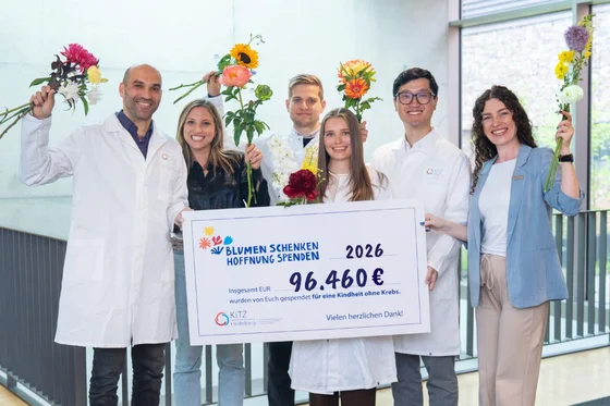 Eine Gruppe von sechs Personen in weißen Laborkitteln steht zusammen und hält Blumen sowie ein großes Schild mit der Aufschrift "96.460€". Darin wird auf eine Spende für eine Kindheit ohne Krebs hingewiesen. Die Atmosphäre wirkt fröhlich und hoffnungsvoll.