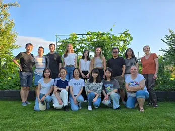 Eine Gruppe von 15 Personen steht und sitzt in einem Garten, umgeben von grünen Pflanzen. Die Menschen lächeln und sind leger gekleidet. Der Himmel ist blau mit wenigen Wolken, und das Gras ist grün. Es scheint ein freundliches, geselliges Treffen zu sein.