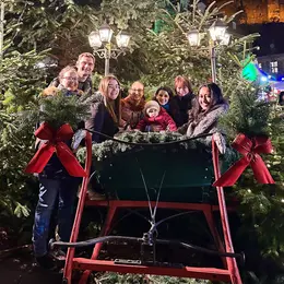 Eine Gruppe von sieben Personen steht um einen festlich geschmückten Schlitten, der mit grünen Tannenzweigen und roten Schleifen verziert ist. Im Hintergrund sind Weihnachtslichter und Tannenbäume sichtbar, was eine festliche Stimmung erzeugt. Das Bild vermittelt ein Gefühl von Gemeinschaft und Feierlichkeit.