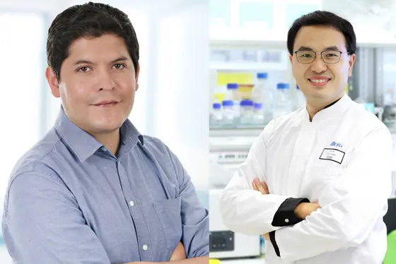 Das Bild zeigt zwei junge Männer, die jeweils lächeln. Der linke Mann trägt ein hellblaues Hemd, während der rechte Mann einen weißen Laborkittel trägt und Brille trägt. Im Hintergrund sind Laborutensilien zu sehen. Das Bild vermittelt die Zusammenarbeit in der Forschung.