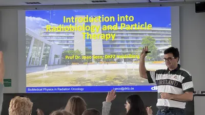 Ein Professor steht vor einer Präsentation mit dem Titel "Einführung in Radioonkologie und Partikeltherapie". Im Hintergrund sieht man eine moderne Gebäudeansicht. Der Professor schwingt eine Hand, während er seine Zuhörer anspricht.