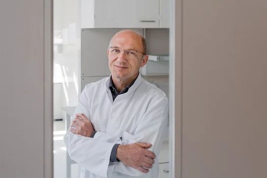 Ein Wissenschaftler in einem weißen Laborkittel steht lächelnd mit verschränkten Armen vor einem hellen, modernen Hintergrund. Hinter ihm sind Regale und eine glänzende Oberfläche zu sehen.
