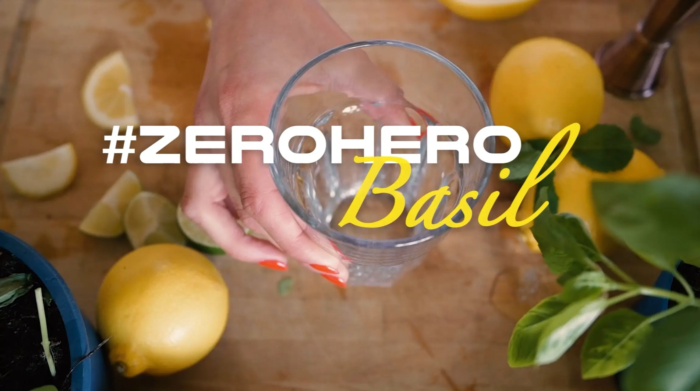Eine Hand hält ein Glas mit Wasser, umgeben von frischen Zitronen und Basilikum. Im Hintergrund sind aufgeschnittene Zitronen und eine Pflanze sichtbar. Der Text "#ZEROHERO Basil" ist prominent platziert und betont das Thema der Frische und natürlichen Zutaten.