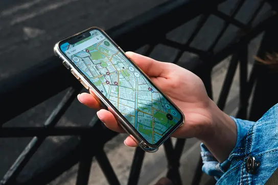 Eine Hand hält ein Smartphone, das eine Kartenanwendung zeigt. Die Karte enthält Straßen, Wege und Punkte von Interesse, die deutlich markiert sind. Der Hintergrund ist unscharf, was den Fokus auf das Gerät und die Karte legt.
