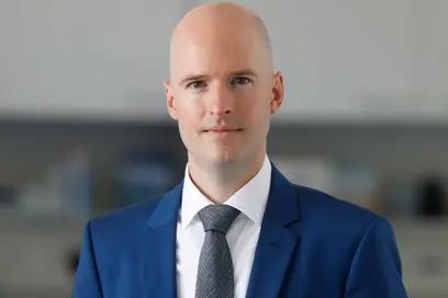 A man with a bald head, wearing a blue suit and a tie, stands in a laboratory setting. He appears confident and professional, with a neutral expression on his face. The background is slightly blurred, focusing attention on him.