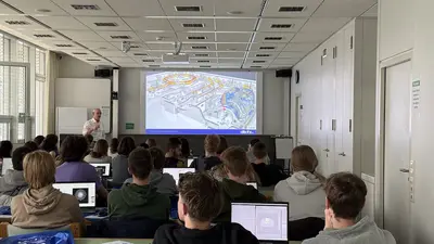 Ein Dozent spricht vor einer Gruppe von Schülern, die an ihren Laptops sitzen. Im Hintergrund ist eine Präsentation mit technischen Diagrammen auf einer Leinwand zu sehen. Die Schüler sind teilweise mit dem Rücken zur Kamera.