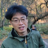 A man with short black hair and glasses smiles while standing outdoors in a wooded area. He is wearing a green jacket and appears to be enjoying the natural surroundings. The background features trees with autumn foliage.