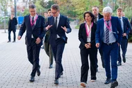 Eine Gruppe von vier Personen geht auf einem Gehweg. Sie tragen elegante Kleidung und haben identische Namensschilder mit rosa Bändern. Die Atmosphäre wirkt informell, und einige Personen scheinen sich zu unterhalten.