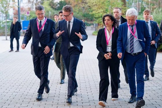 Eine Gruppe von vier Personen geht auf einem Gehweg. Sie tragen elegante Kleidung und haben identische Namensschilder mit rosa Bändern. Die Atmosphäre wirkt informell, und einige Personen scheinen sich zu unterhalten.