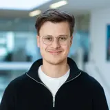 Ein junger Mann mit Kurzhaarschnitt und Brille lächelt in die Kamera. Er trägt einen schwarzen Pullover und ein weißes T-Shirt. Der Hintergrund zeigt eine helle, moderne Innenarchitektur.