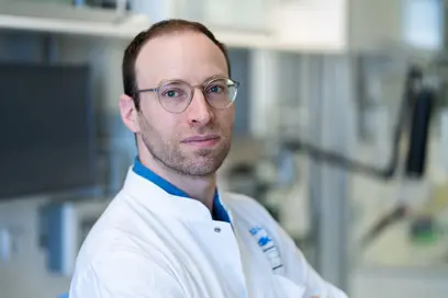 Ein Mann in einem weißen Laborkittel steht in einem modernen Labor. Er trägt eine Brille und schaut direkt in die Kamera. Der Hintergrund zeigt Geräte und Laborelemente, die auf eine wissenschaftliche Umgebung hinweisen.