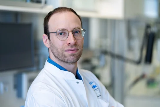 Ein Mann in einem weißen Laborkittel steht in einem modernen Labor. Er trägt eine Brille und schaut direkt in die Kamera. Der Hintergrund zeigt Geräte und Laborelemente, die auf eine wissenschaftliche Umgebung hinweisen.