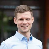 Ein junger Mann mit kurzem, blondem Haar lächelt in die Kamera. Er trägt ein hellblaues Hemd und steht vor einem unscharfen, modernen Hintergrund, der Pflanzen und Gebäude andeutet.