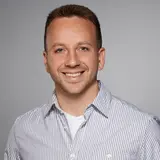 Porträt eines lächelnden Mannes mit kurzen Haaren, der ein gestreiftes Hemd trägt. Der Hintergrund ist grau und neutral. Der Mann strahlt Freundlichkeit und Selbstbewusstsein aus.