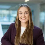 Eine lächelnde Frau mit langen, glatten Haaren steht in einem modernen Innenraum. Sie trägt einen dunkelroten Blazer über einem weißen Oberteil und hat ihre Hand lässig an einem Geländer abgelegt. Ihr Gesicht zeigt Freude und Selbstbewusstsein.