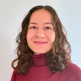 Porträt einer lächelnden Frau mit lockigem Haar, die einen roten Pullover trägt. Der Hintergrund ist hell und neutral.