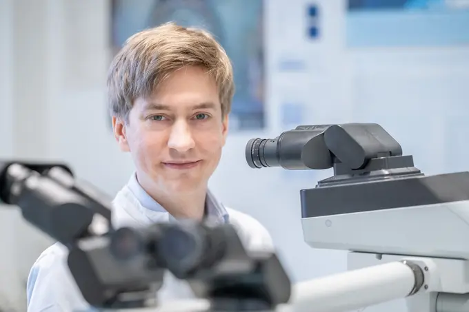 Ein junger Mann mit blonden Haaren lächelt freundlich in die Kamera. Im Hintergrund sind Mikroskope sichtbar, was auf eine wissenschaftliche oder medizinische Umgebung hinweist.