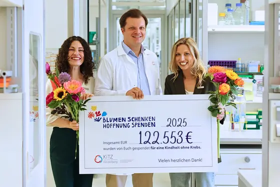 Drei lächelnde Personen halten in einem Forschungslabor einen großen symbolischen Spendenscheck mit der Aufschrift „Blumen schenken, Hoffnung spenden – 122.553 € für eine Kindheit ohne Krebs“. Zwei Frauen halten bunte Blumensträuße.