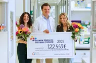 Drei lächelnde Personen halten in einem Forschungslabor einen großen symbolischen Spendenscheck mit der Aufschrift „Blumen schenken, Hoffnung spenden – 122.553 € für eine Kindheit ohne Krebs“. Zwei Frauen halten bunte Blumensträuße.
