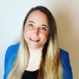 Eine lächelnde Frau mit langen, glatten Haaren steht vor einer hellen Wand. Sie trägt ein schwarzes Oberteil und eine blaue Jacke. Ihr freundlicher Ausdruck vermittelt Offenheit und Zugänglichkeit.