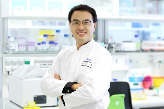 Ein Wissenschaftler in einem Labor steht mit verschränkten Armen lächelnd vor einem Tisch mit verschiedenen Laborausrüstungen und Chemikalien. Er trägt einen weißen Laborkittel und eine Schutzbrille. Der Hintergrund zeigt Regale mit bunten Behältern.